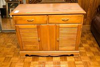 Front view of wooden sideboard showing two drawers with brass handles, three doors (two louvered) below, on parquet floor.