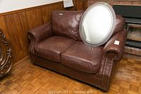 Full view of brown leather love seat with rolled arms and brass nail head trim; oval mirror leaning against back cushion