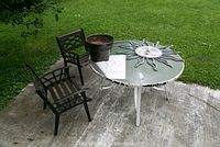 Outdoor glass top table with metal base, two chairs, pots, and decorative sun ornament arranged on patio concrete area with grass background