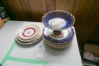 Stack of five floral and gold-trimmed plates with visible crazing, placed beside stacked blue and gold footed bowl and additional plates.
