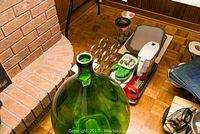 Photo showing large green glass demijohn with narrow neck and bulbous body, metal wine rack, wine cooler, oven mitts, corkscrew, placemats, and kitchen towels arranged on hardwood floor beside brick hearth.