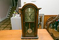 Full view of the wooden mantel or wall clock on a table, showcasing its curved top, glass front, and visible pendulum and weights inside.