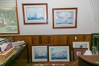 Photo showing all four framed nautical prints arranged on the wall and floor, each in brown wooden frame with white and grey double matte, depicting different maritime scenes with sailing ships.