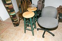 Front view of two wooden stools with round natural wood seats, green legs, one stool has repaired seat, a wooden chair with a curved back, and a gray office chair with armrests and wheels on a tiled floor.