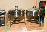 Two round stainless steel chafing dishes with lids on a wooden shelf.