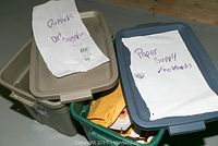 Two plastic storage bins with handwritten labels indicating contents: 'Puppets Art Supplies' and 'Paper Supply no notebooks'. Bins are beige and grey with lids.