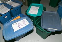 Blue and green plastic bins labeled with hand-written notes containing paper sheets and supplies.