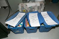 Three blue plastic bins with papers taped on top labeling them as Teacher's play cash register, Language resources ages 6-12, and Environmental resources.