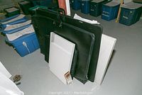 View showing two black portfolio cases standing upright with handles and clasps, three white canvas boards leaning against them, and a small box of art supplies in front.