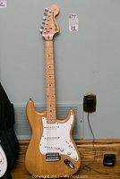 Full view of natural finish guitar leaning against wall with gig bag