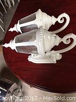 Pair of white outdoor coach-style wall lamps shown side by side on table, clear glass panels, curved arms, hexagonal lantern shape.