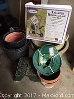 Photo showing unopened box of Claber hose reel cart, green garden hose coiled with attached nozzle, several planters and small green step stool.