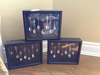 Three black-framed shadow boxes with seven collector spoons displayed in each, arranged on the floor against a wall.