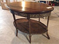 Oval parlour table showing wood top, wicker apron and legs, and lower wood shelf on scalloped edge base.