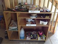 Handmade wooden rolling storage bench with multiple shelves holding various items including cleaning supplies, hardware boxes, electrical outlet covers, and tape roll.