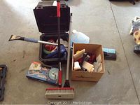 Overall view of lot items including black plastic case with car polisher, open cardboard box with paint supplies, broom with red handle, and blue jug.
