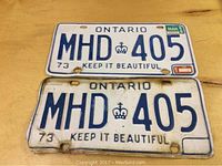Two Ontario license plates dated 1973 with registration MHD 405. One plate bent and rusted, the other with original validation stickers.