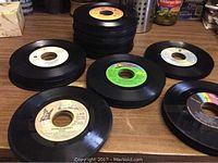 Stack and scattered arrangement of multiple 45 RPM vinyl records on wooden table, showing variety of label colors and wear.