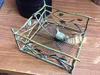Front angled view of square metal napkin holder showing coiled circular base with small bird figurine and branch and leaf sides.