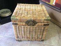 Full front view of a square wicker chest with a hinged lid, natural coloration, metal corner supports and front lock.