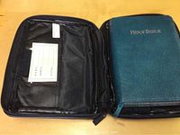 Teal leather bound Holy Bible inside a black leather zippered carry case, showing the outer cover of the Bible and interior pocket on the case.