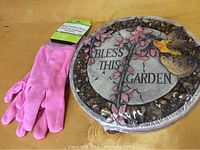 Top-down view showing the pink gardening gloves next to the round garden stone with floral and bird design and inscription 'Bless This Garden'.