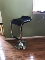 One of the two adjustable swivel bar stools with a black curved seat and chrome base, positioned on a wooden floor near a green wall and window.