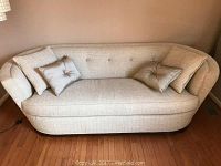 Beige curved upholstered sofa with four decorative pillows placed on wooden floor against a wall. Shows button tufting on backrest and slight wear on wooden base.