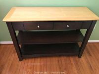 Front view of a wooden server with a light wood top contrasting with the dark wood frame, two drawers with metal knobs under the top surface, and two shelves below for storage display.