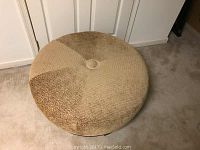 Photo of a large round ottoman placed on carpeted floor, showing top view of the patchwork fabric upholstery with shades of brown and beige and central button tuft.