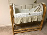 Infant rocking cradle with wooden frame, fabric lining, beige gingham skirt, mesh canopy, and attached music box.