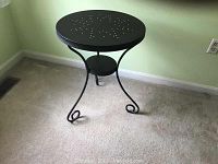 Photo of a small black metal table with round perforated top and decorative curled legs showing the whole item on carpeted floor.