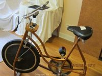 Full view of vintage Schwinn exercise bicycle showing mustard gold frame, large black front wheel cover, pedals, two seats, and handlebars with bell.