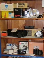 Photograph of multiple vintage cameras, lenses, flash units, and boxed accessories arranged on wooden shelving.