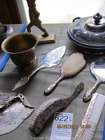 Photo shows assorted silver plated vanity items including brushes, dust pan, and cake server displayed on a table with a small brass urn and brass figurine in background.