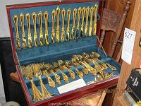 Open wooden box showing full gold plated flatware service for 16 with various serving pieces, arranged inside.