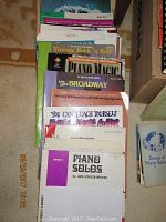 Photo showing a stack of various sheet music books including vintage rock and roll, Broadway collections, piano solos, and guitar instruction books.