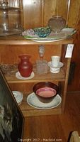 Photo showing various vintage glassware pieces on a small wooden bookcase, including a glass cake dish with lid, two platters, salad bowl, covered casserole, light fixture, red vase, cut glass bottle, and teacups.