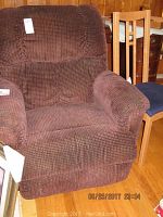 Brown fabric reclining chair and wooden dining chair with blue seat shown side-by-side against wooden panel wall.