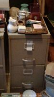 Photo of beige 2 drawer metal file cabinet with slight rust and dents, items on top include ceramic pitcher, sugar bowl with lid, small containers, and trays