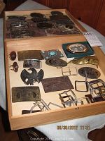 Photo showing a wooden drawer with 20 assorted belt buckles and belt accessories arranged in rows, featuring various metallic finishes and shapes including oval, rectangular, and decorative designs with floral, eagle, and letter motifs.