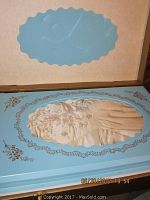 Original blue storage box with raised window displaying a portion of the cream wedding dress with beading and gathered fabric, ornate floral decoration around window, 'Bianchi' scripted inside.