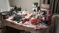 Table with assorted holiday and Christmas items including tins, garlands, lights, candles, wrapping paper, and Halloween pumpkin.