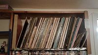 Photo of a wooden shelf packed tightly with numerous vinyl record album covers, mostly paper sleeves showing wear from age and use.