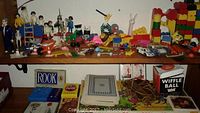 Shelf top displays vintage Playmobil figures of various types including historical and modern characters, Fisher Price Little People toys, miniature cars, and colorful wooden stacking blocks. Various small toys and accessories are scattered.