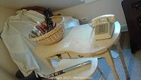 Wooden vanity table with one drawer and a matching wooden stool covered by a light cushion. Basket on top containing assorted Avon cosmetic bottles and tubes. Tan rotary dial phone placed on the vanity surface.