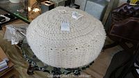 Full view of the cream-colored chunky knit pouf showing woven lambswool texture and round shape with a tag attached providing lot number.