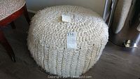 Full view of round, hand knitted pouf in thick natural wool yarn, showing texture and size relative to surrounding furniture.