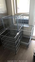 Three assembled metal storage shelving frames with wire basket shelves stacked inside. Each frame has four baskets, 12 baskets total. The frames are rectangular and made of metal wire mesh.