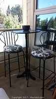 Full view of black iron bar-height table with round top and two matching black metal chairs with black and white patterned cushions.